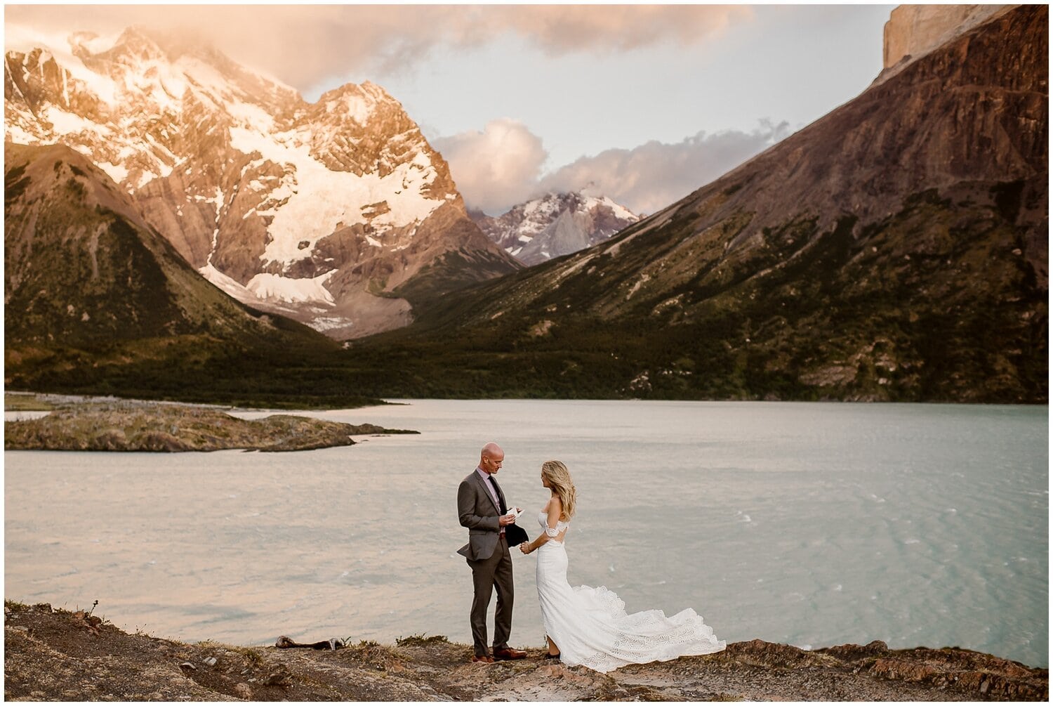 Groom reads his vows to bride in front of a blue alpine lake, with snow covered mountains behind them. 