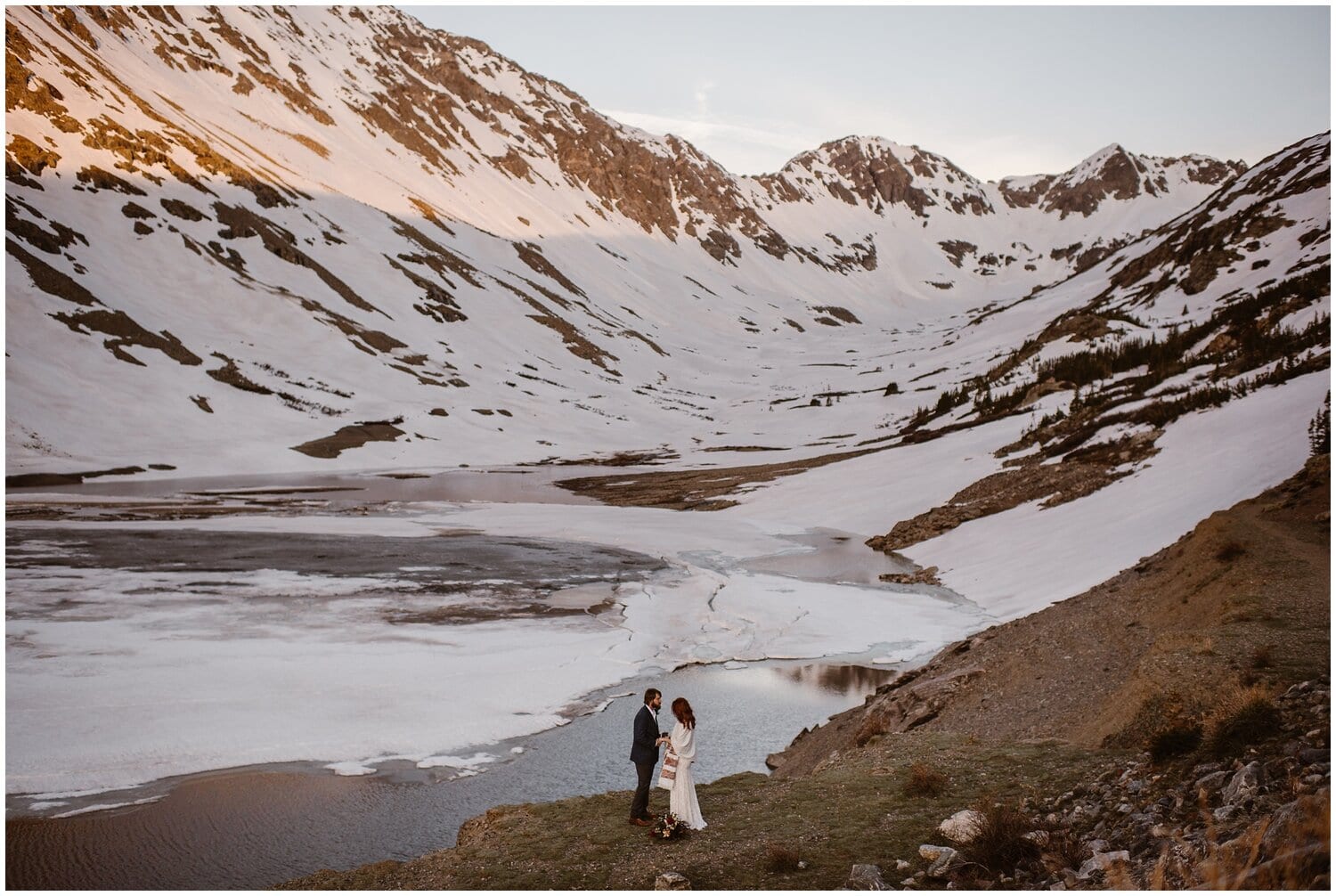 Bride and groom stand in front of a frozen alpine lake. 