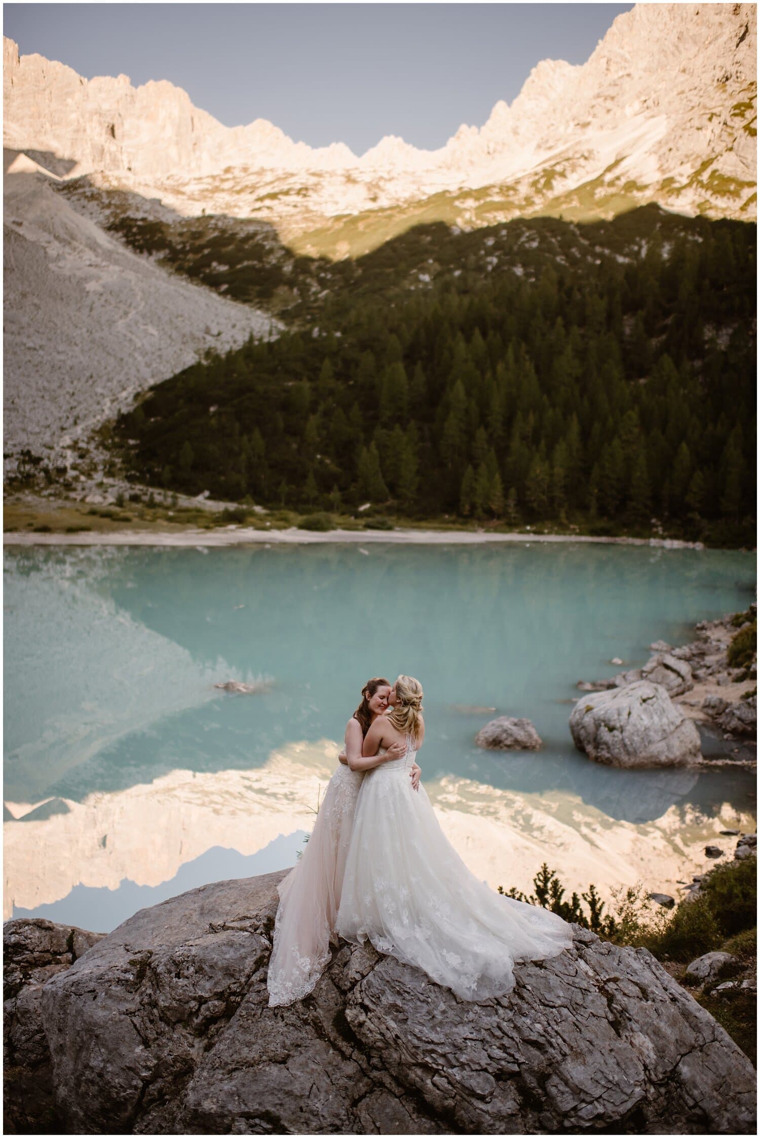 Two brides hug on a rock in front of a blue alpine lake. 