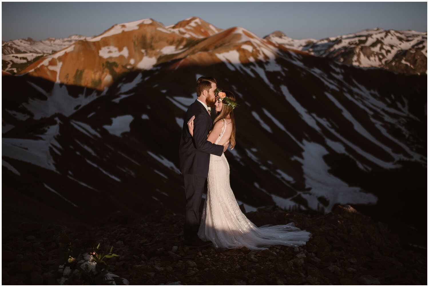 Bride and groom embrace, as groom kisses bride on the cheek. Snow on the mountains behind them. 
