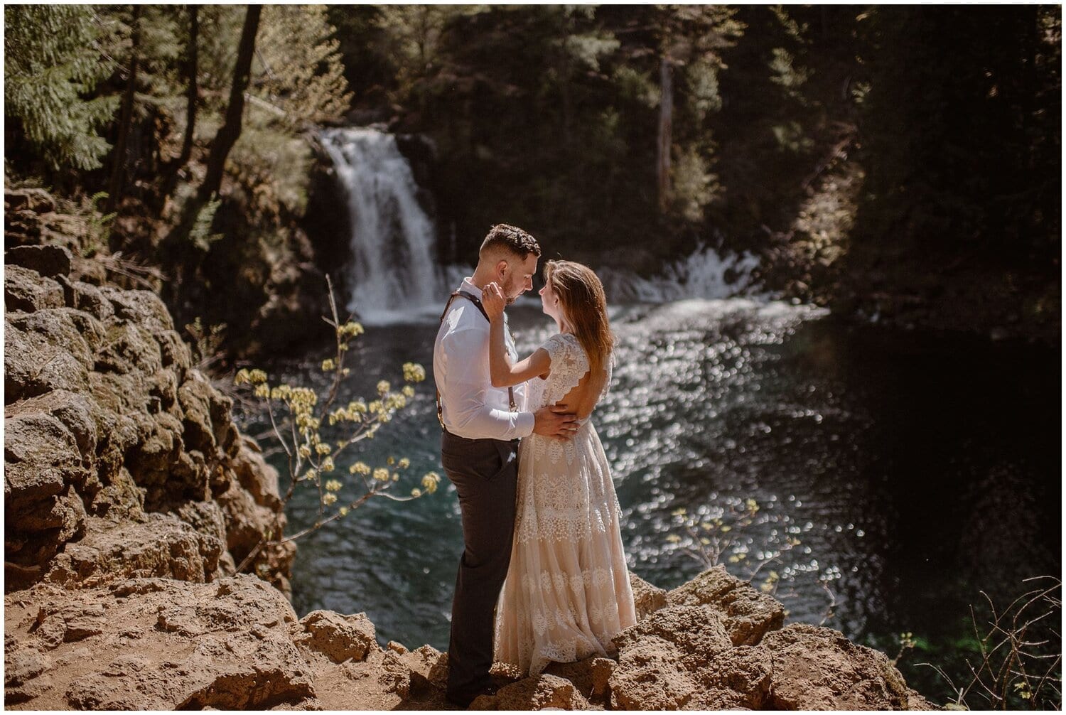 Bride and groom embrace, with waterfall behind them. 