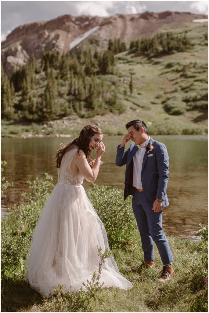 Bride and groom both wipe away tears, with lake behind them. 