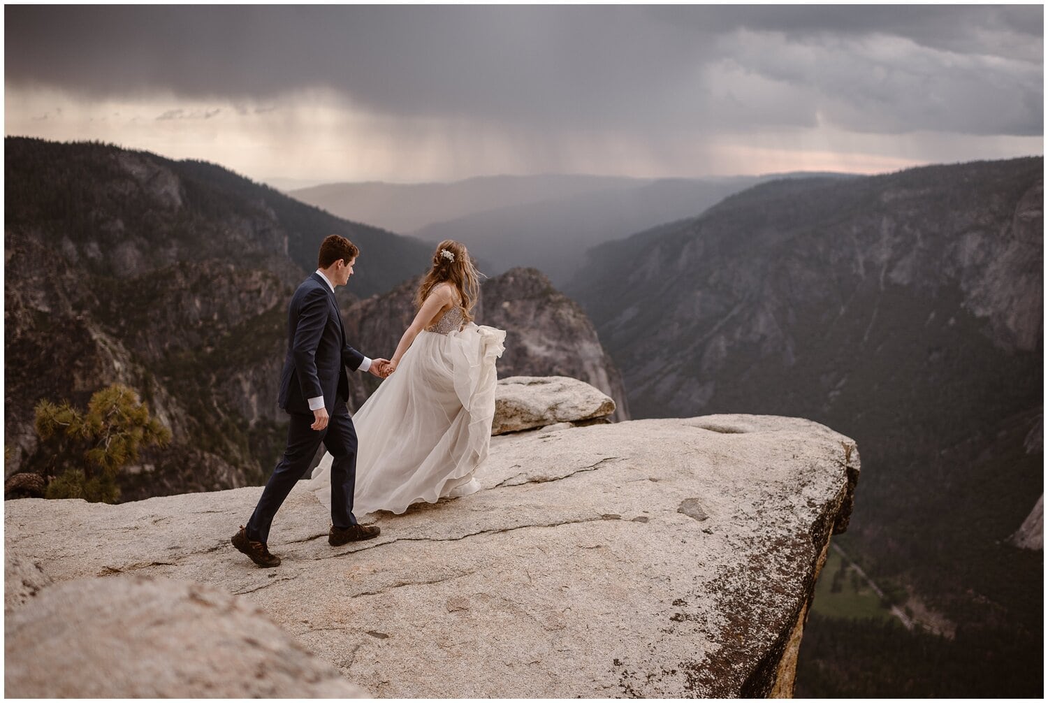 Bride and groom walk on cliff together that overlooks mountain landscape. 