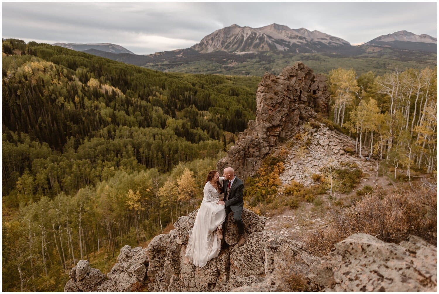 Bride and groom sit on rock, smiling, with green forest behind them. 