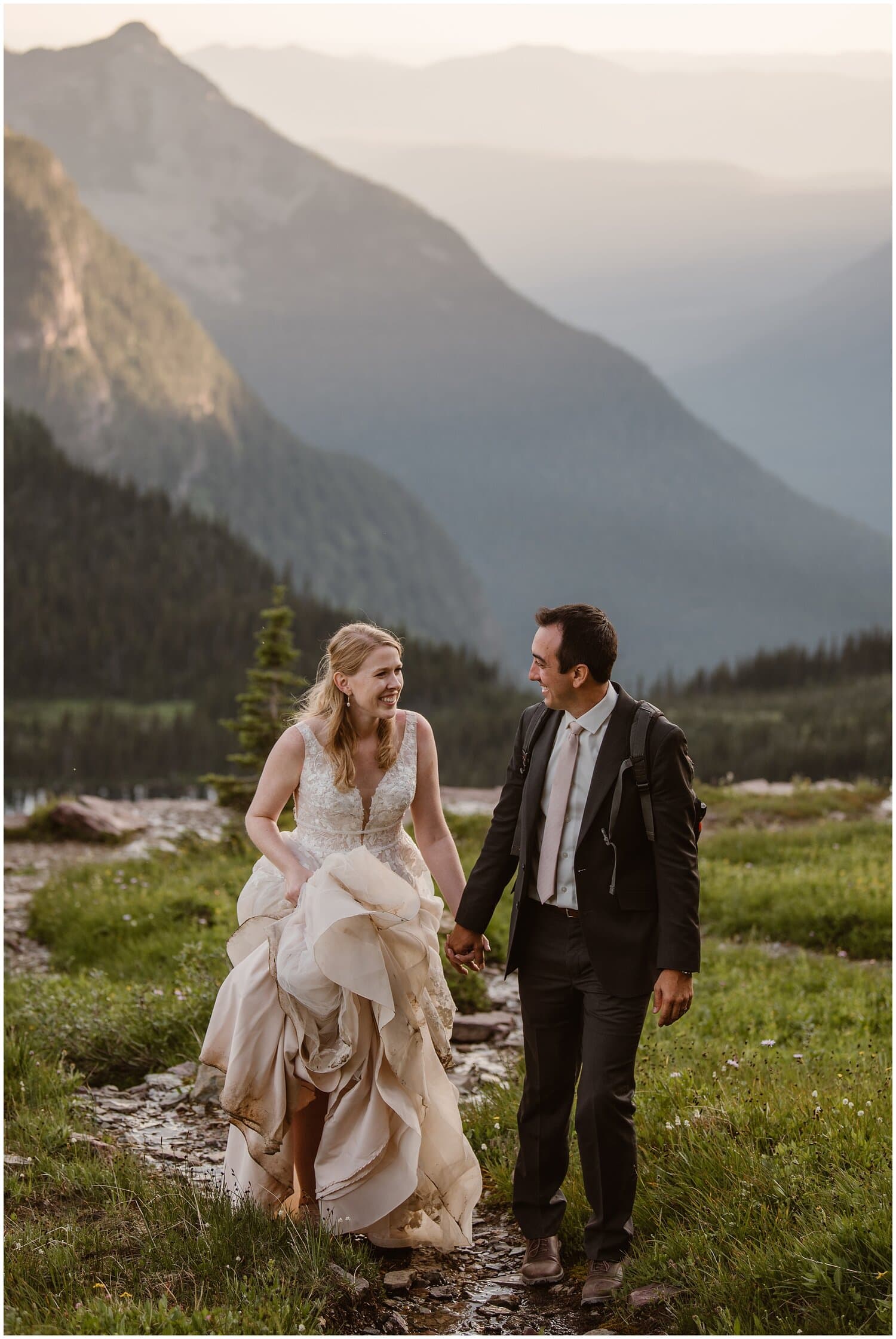 Bride and groom hold hands and smile at each other, while walking on a trail. 