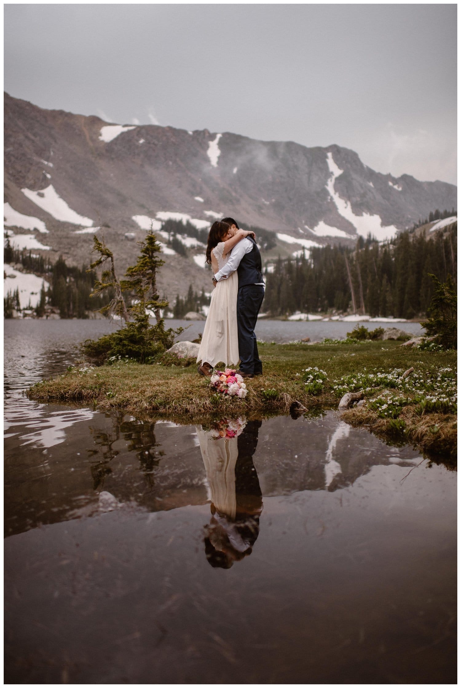 Bride and groom embrace in front of a high alpine lake near Boulder, Colorado. There is a forest and snow-capped mountains in the background. 