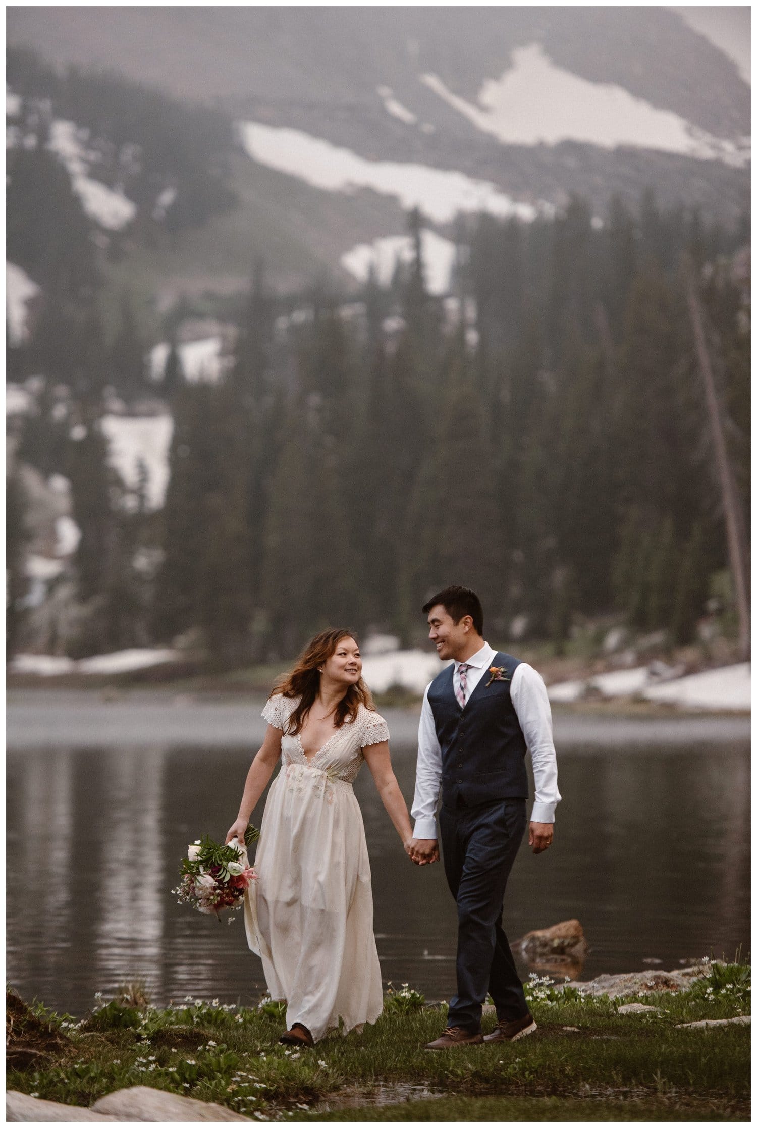 Bride and groom hold hands and smile at each other while standing in front of a high alpine lake near Boulder, Colorado. 