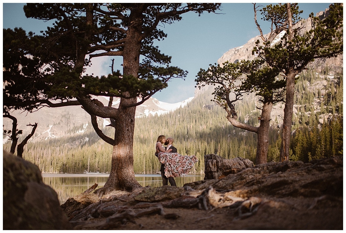 Groom lifts up bride and they share a kiss, with alpine lake, trees, and mountains in the background. 