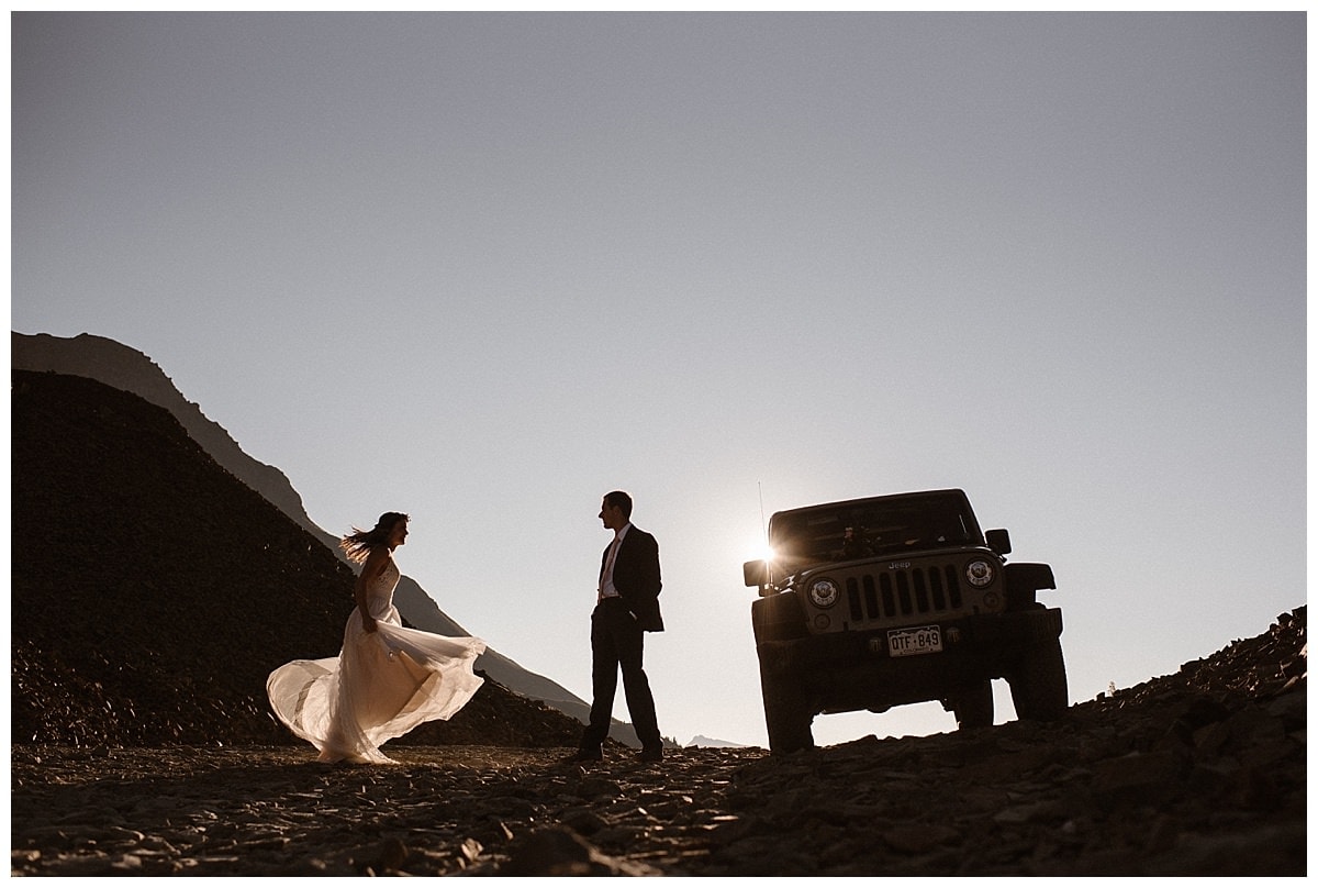 Bride and groom standing next to Jeep at Ophir Pass, in Colorado. The sun is shining behind them as bride holds up her white dress. 