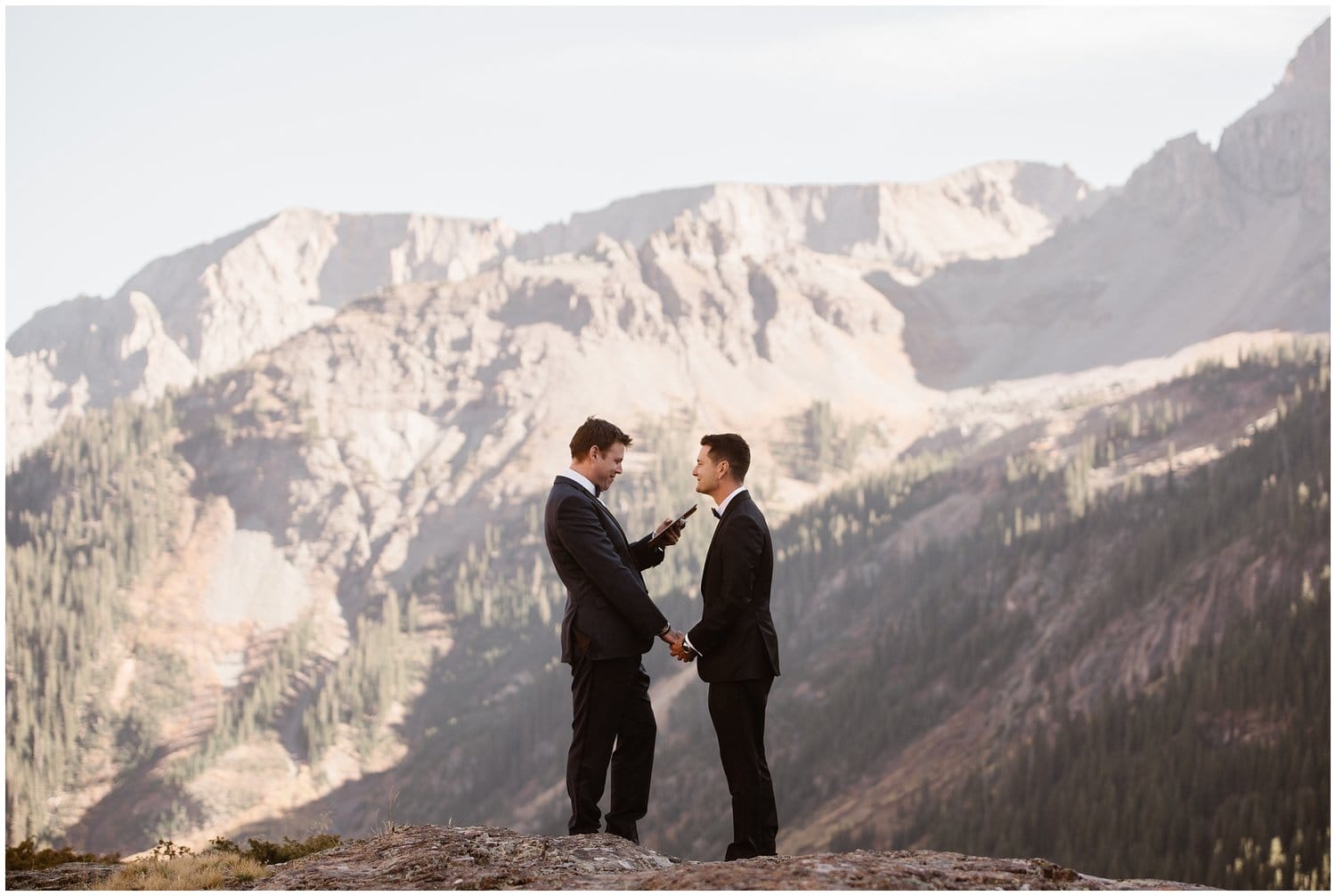Two grooms read their vows, with mountains in the background, during their all-day elopement in Colorado. 