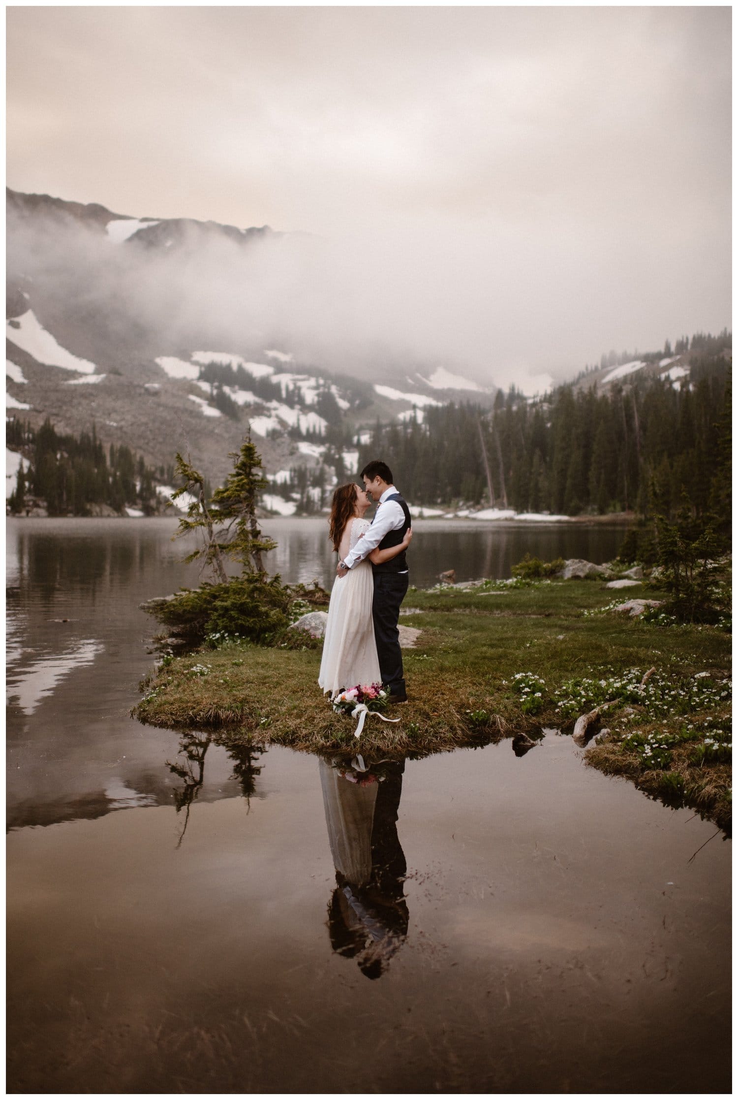 Bride and groom embrace while smiling at each other and standing in front of a high alpine lake, near Boulder, Colorado. There are trees and snow-capped mountains in the background.