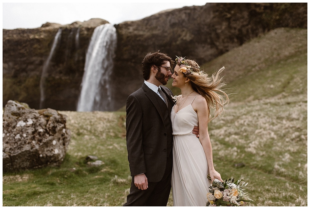 Bride and groom wrap their arms around each other, smiling, in front of a waterfall in Iceland. 