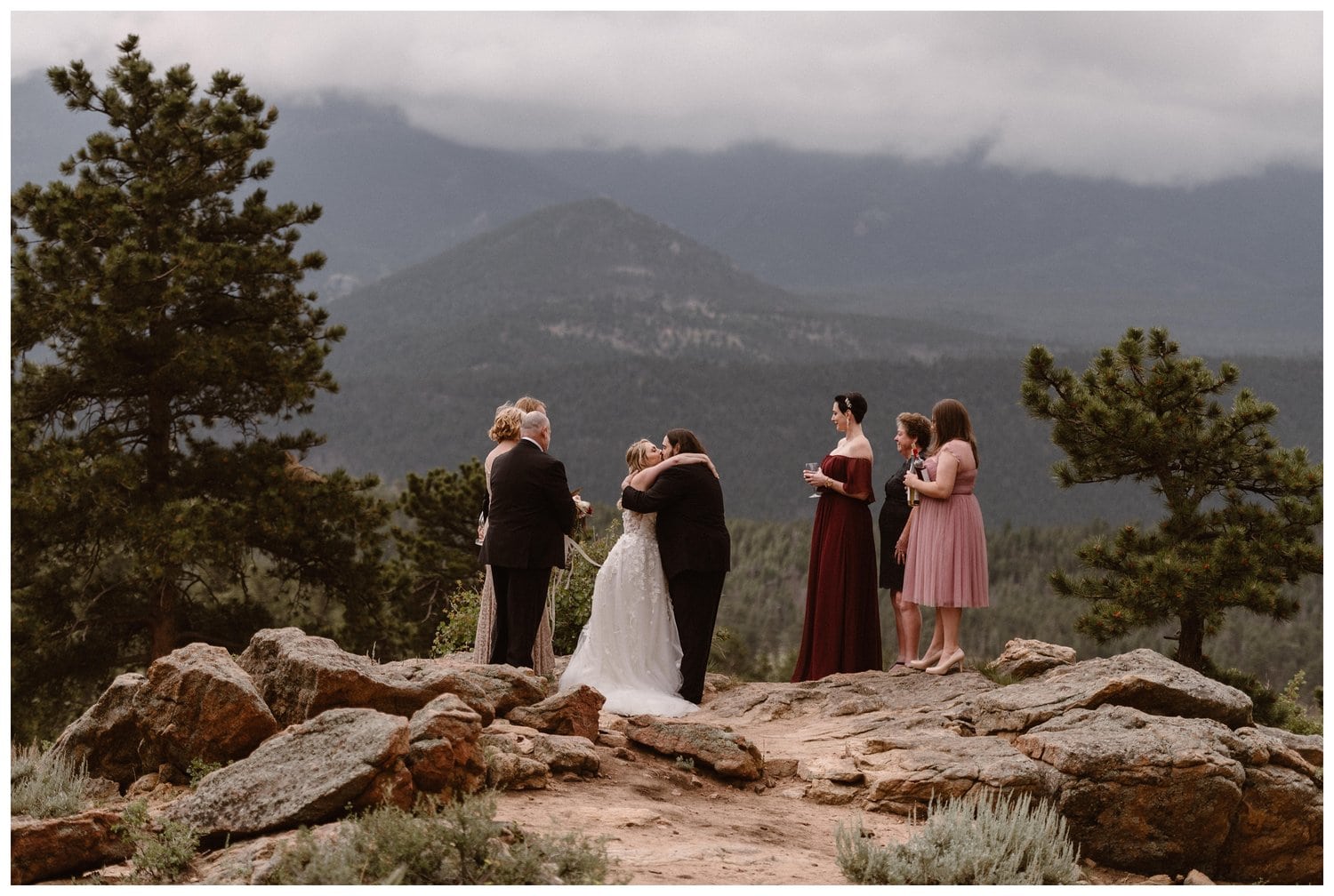Bride and groom kiss during their elopement ceremony, surrounded by friends and family. There are mountains and trees in the background at 3M Curve in Rocky Mountain National Park, Colorado. 