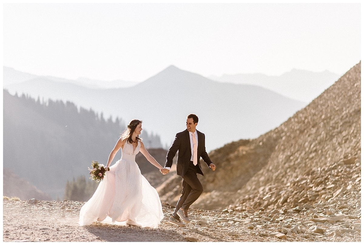 Bride and groom hold hands and walk on trail at Ophir Pass, in Colorado.