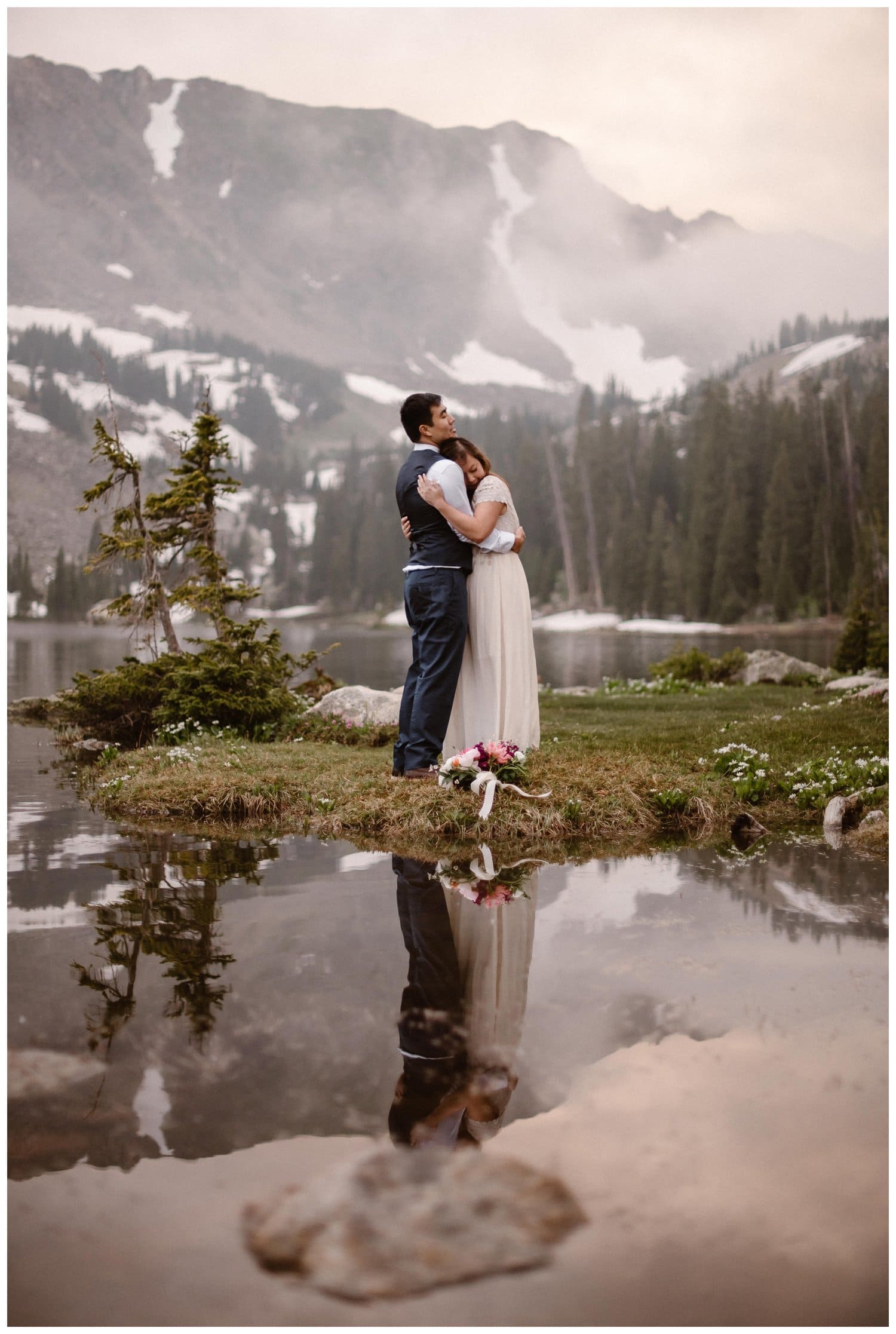 Bride and groom embrace while standing in front of a high alpine lake at sunset, near Boulder, Colorado. 