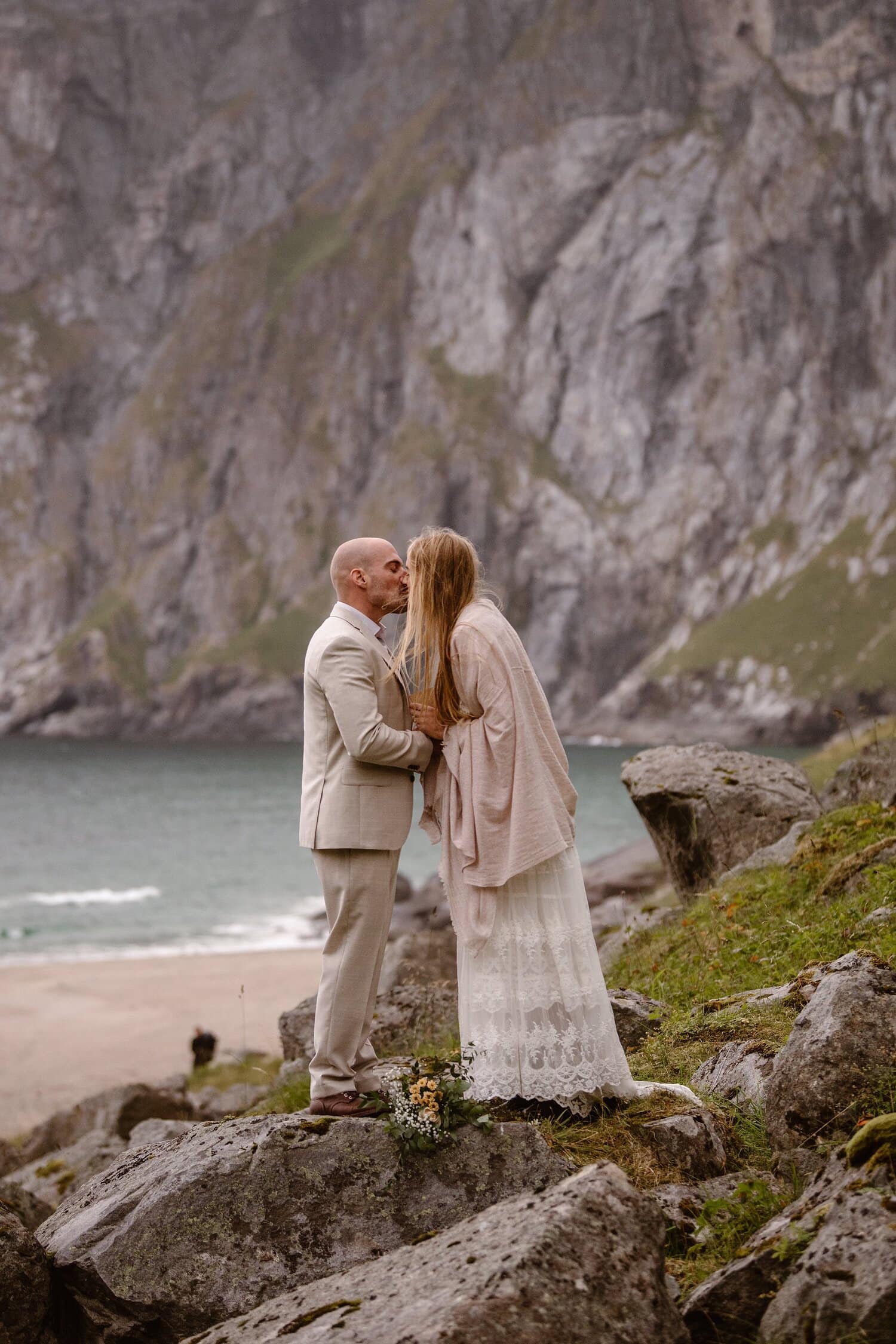 Bride and groom kiss during their intimate elopement ceremony on a beach in Norway. There are mountains in the background.