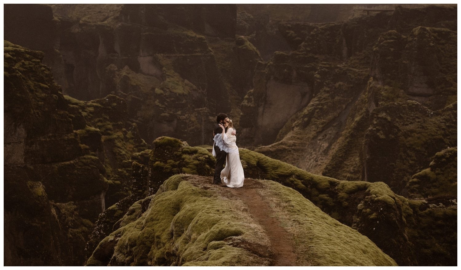 Bride and groom embrace while standing on a top of a mossy green canyon on their elopement day. 
