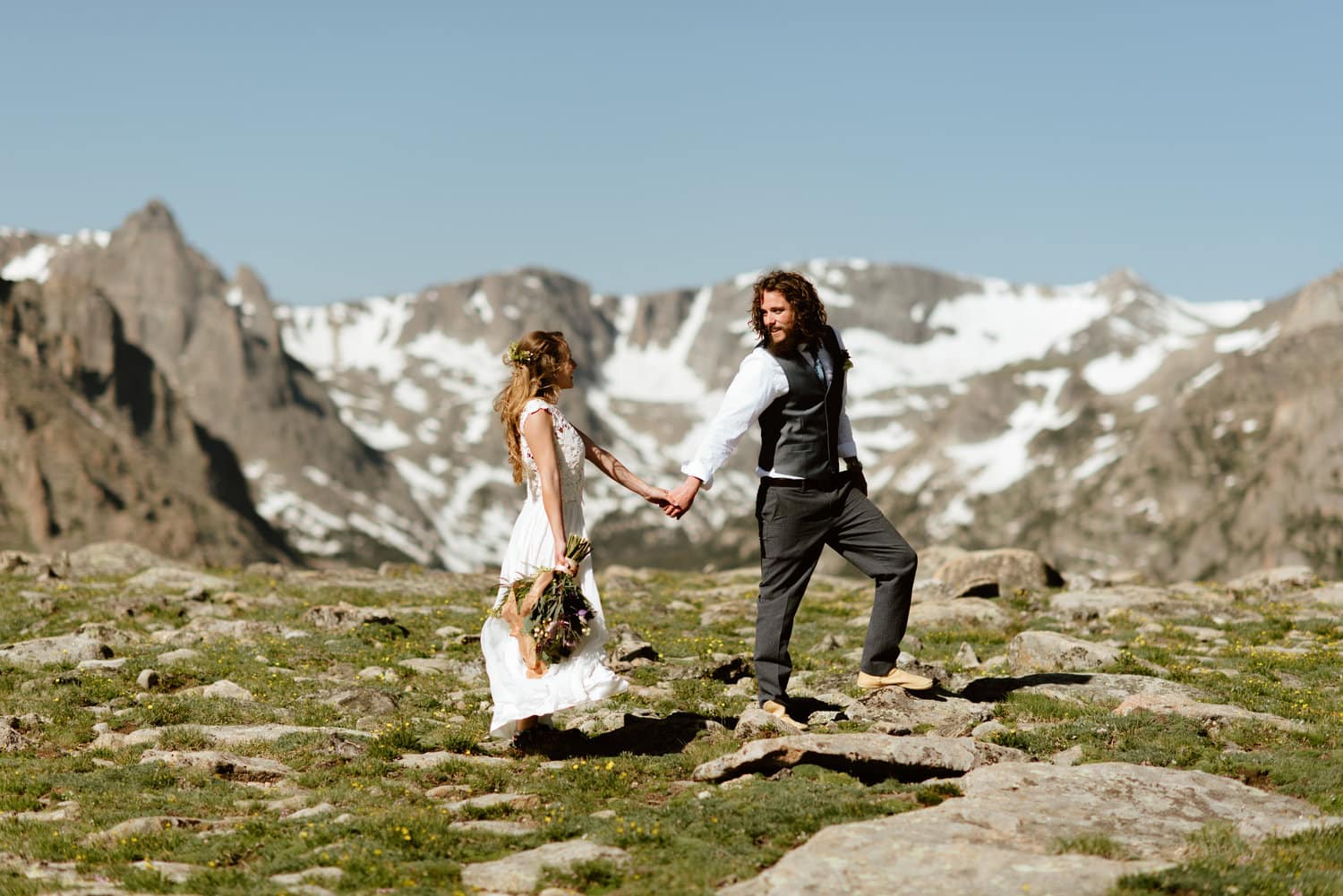 Bride and groom hold hands while smiling at each other and walking around Trail Ridge Road, Colorado. There are snow-capped mountains in the background. 