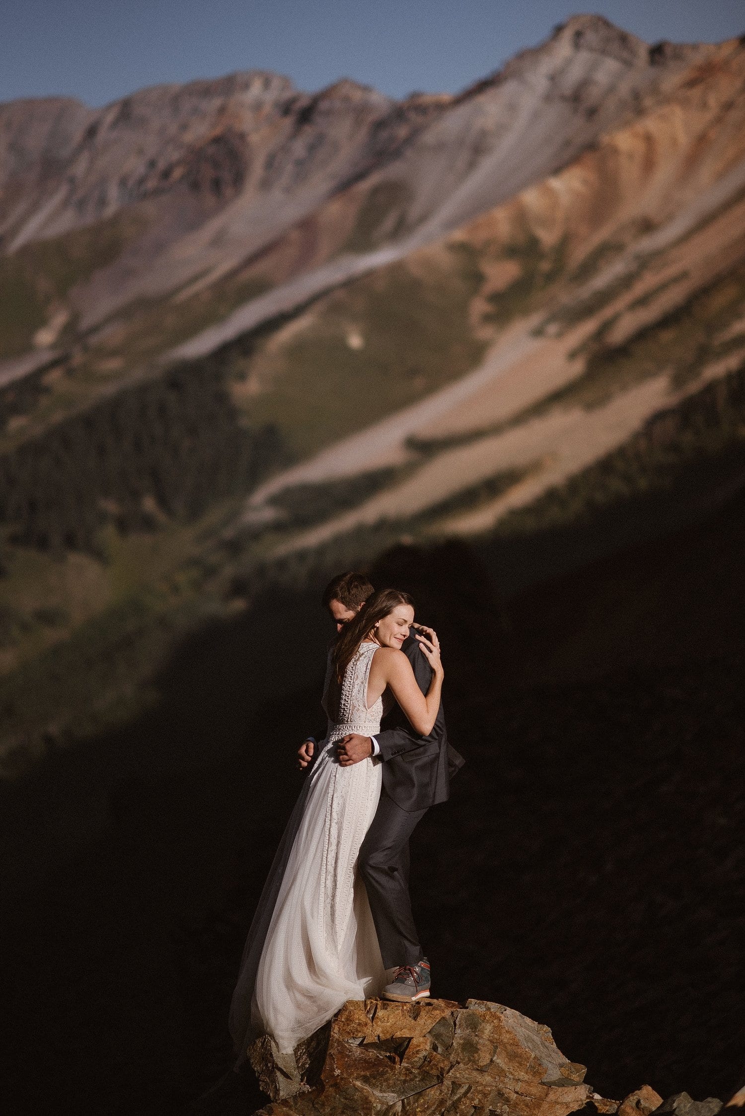 Bride and groom embrace while standing on a rock. There are mountains in the background. 