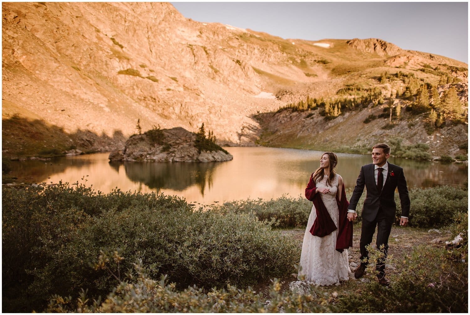 Bride and groom look off in the distance while holding hands in front of alpine lake. 