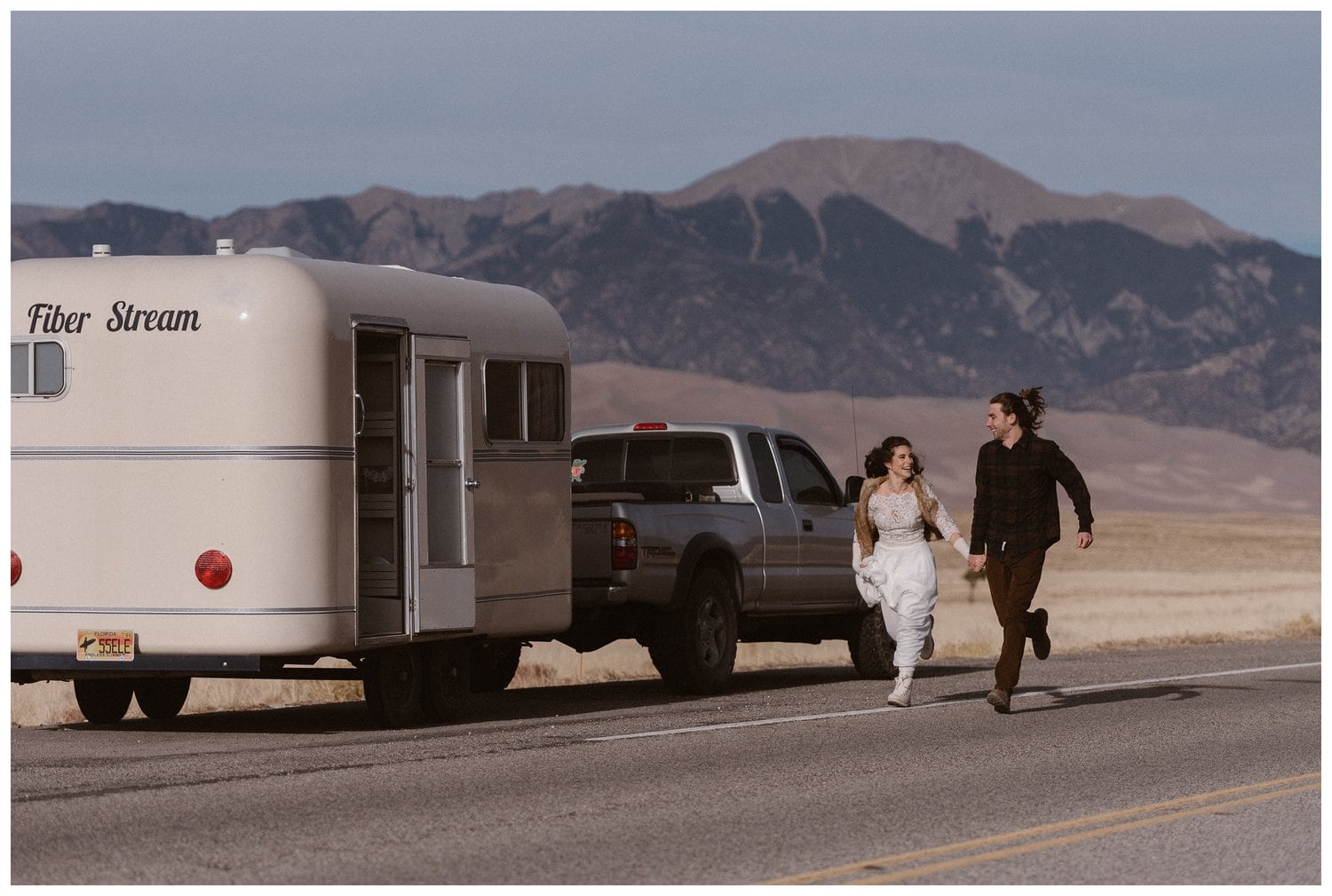 Bride and groom laughing and running, hand-in-hand, next to camper in Colorado. 