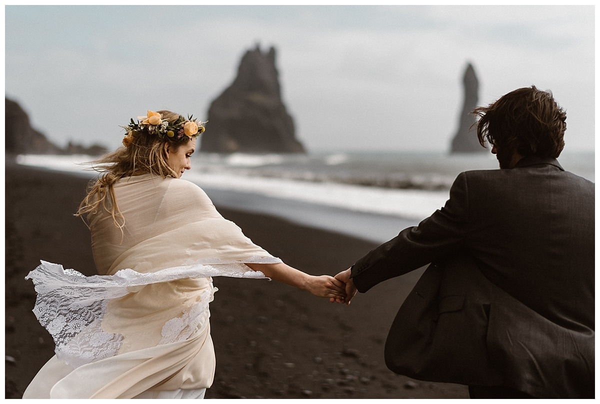 Bride and groom hold hands and walk on a black sand  beach in Iceland. 