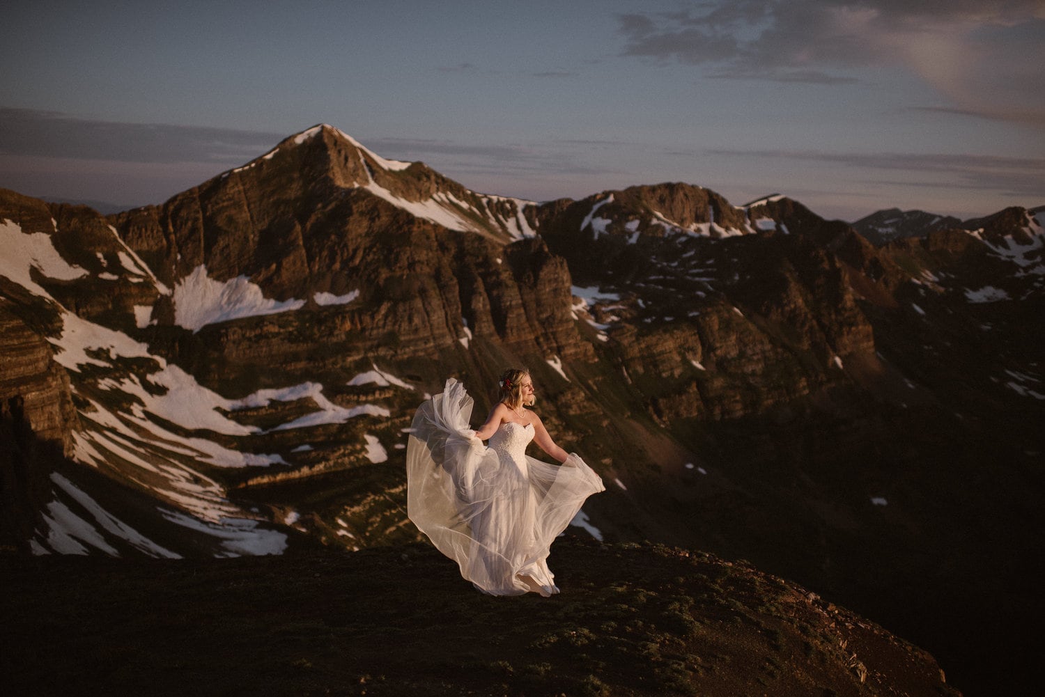 Bride standing on a mountain and holding up the skirt of her white dress, which is flowing in the wind. There are snow-capped mountains in the background. 