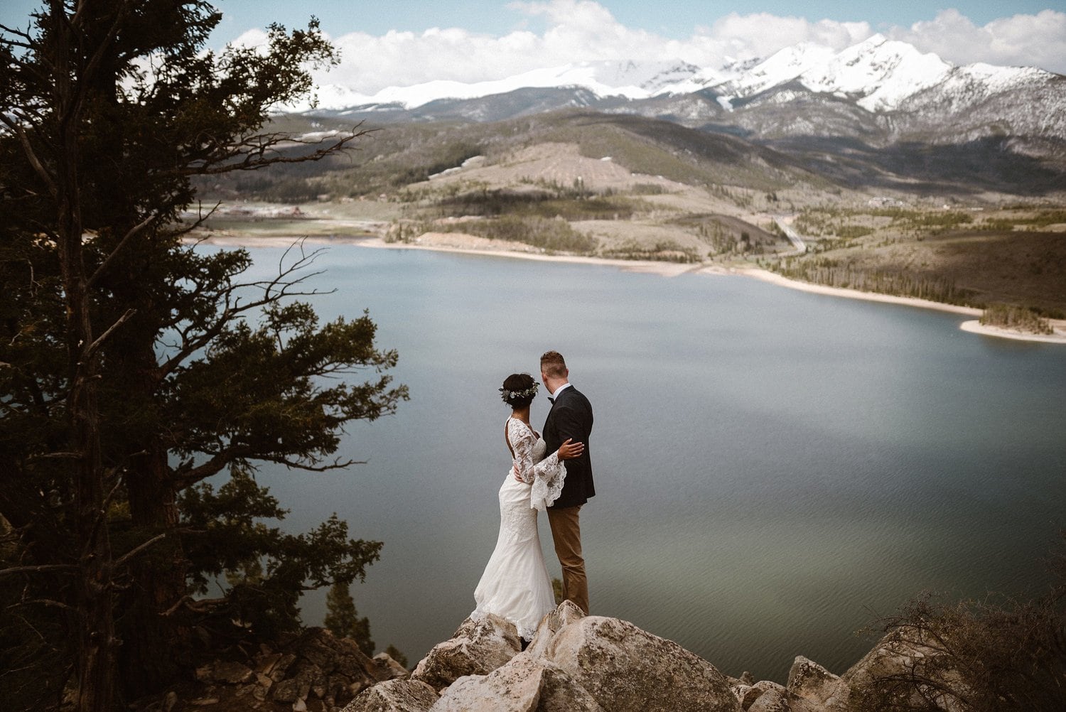 Bride and groom embrace while standing on rocks that overlook at lake at Loveland Pass, in Colorado. 