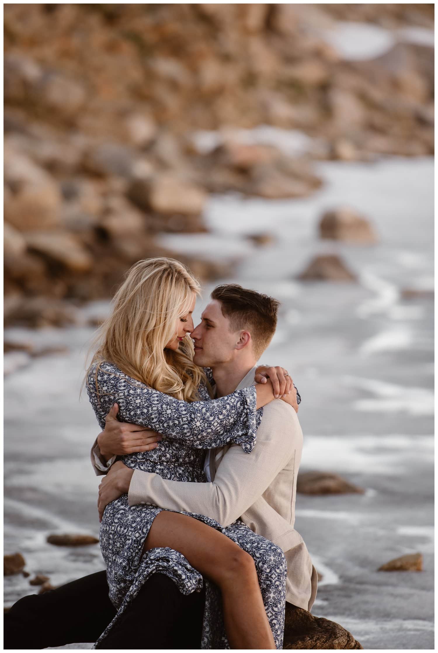Couple embraces while sitting on a rock at Mt. Evans in Colorado. There is a frozen lake behind them. 