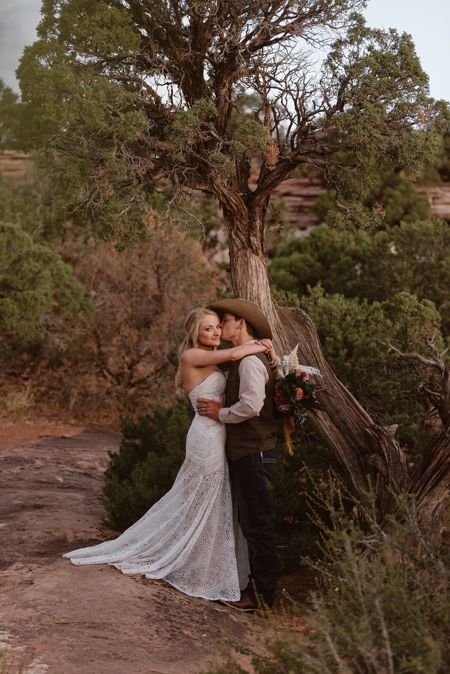 Groom kisses bride on cheek underneath a tree at Colorado National Monument. 