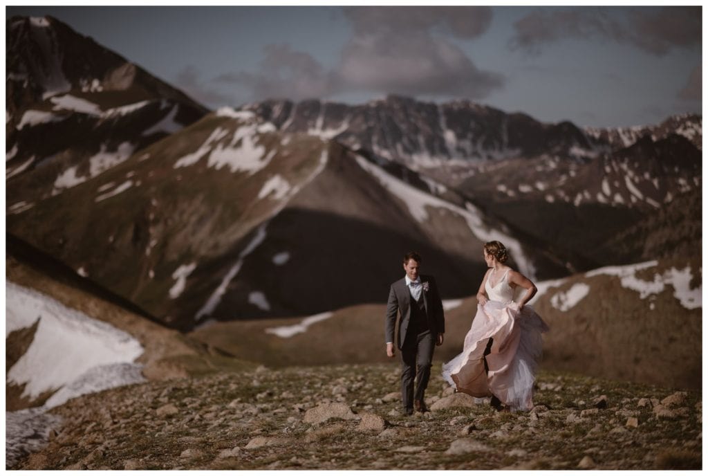 Bride and groom walk along a mountain ridge together during a sunrise hiking elopement near Aspen, Colorado. Bride is holding up the skirt of her dress, which is flowing behind her. 