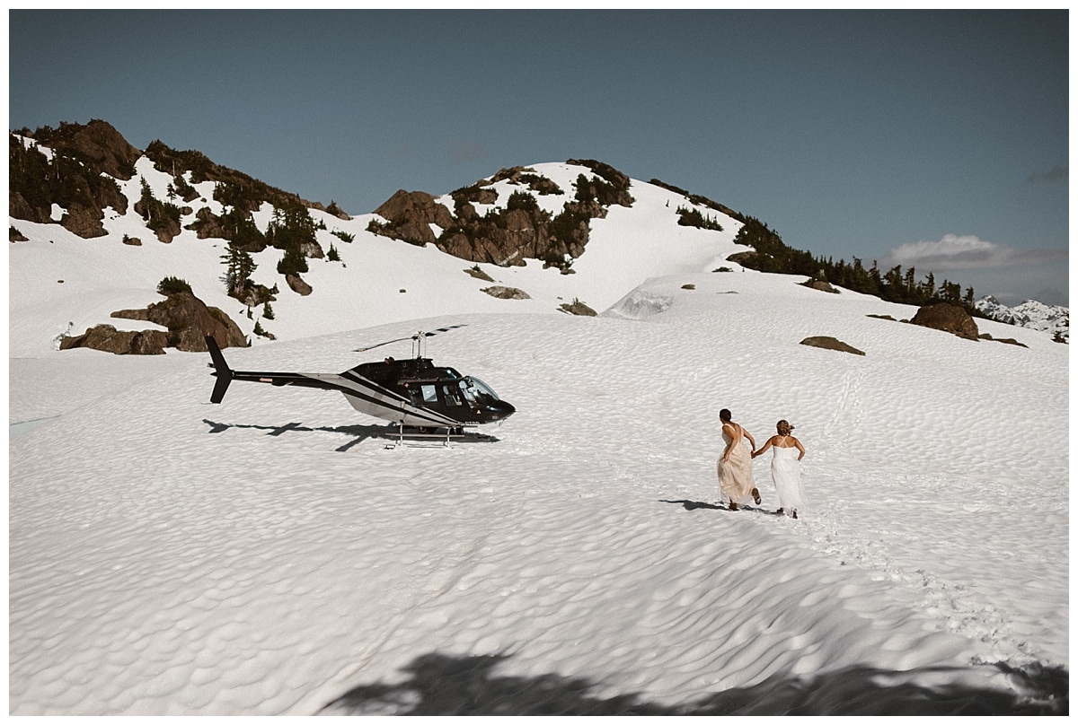 Two brides run through the snow on a glacial mountain in Tofino, Canada. There is snow covering the ground and a helicopter in the background. 