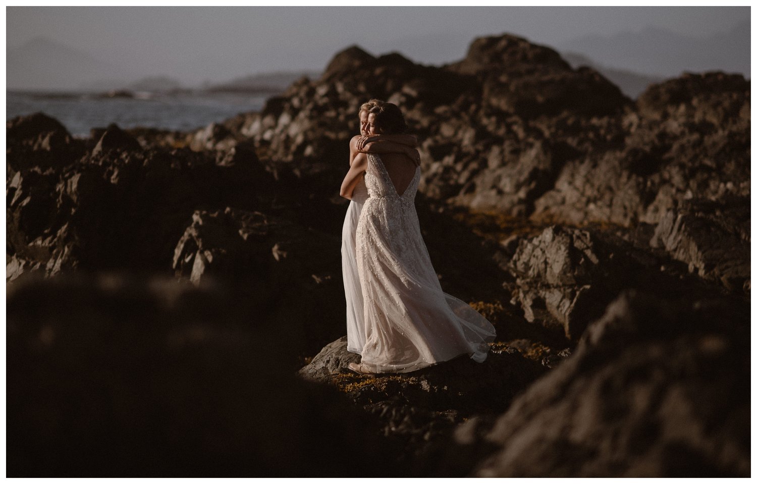 Two brides hug while standing on coastal rocks, on their elopement day. 