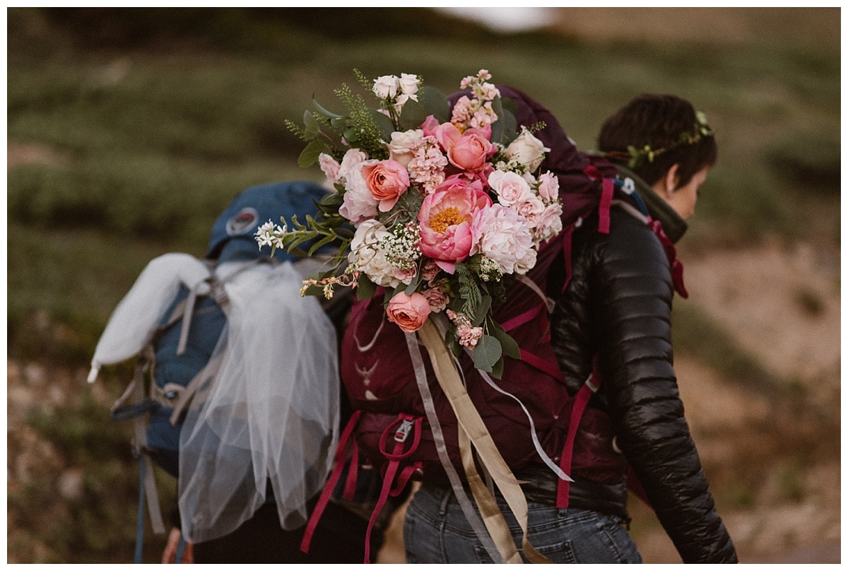 Two brides hiking in Winter Park, Colorado. They are wearing backpacks, one has a bouquet tucked in it with pink roses and peonies. 