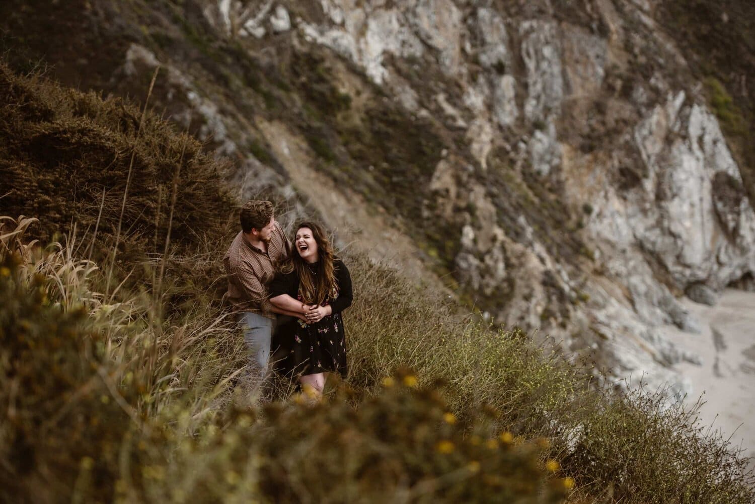 Bride and groom laugh together surrounded by tall grass, next to the beach, in Big Sur, California.