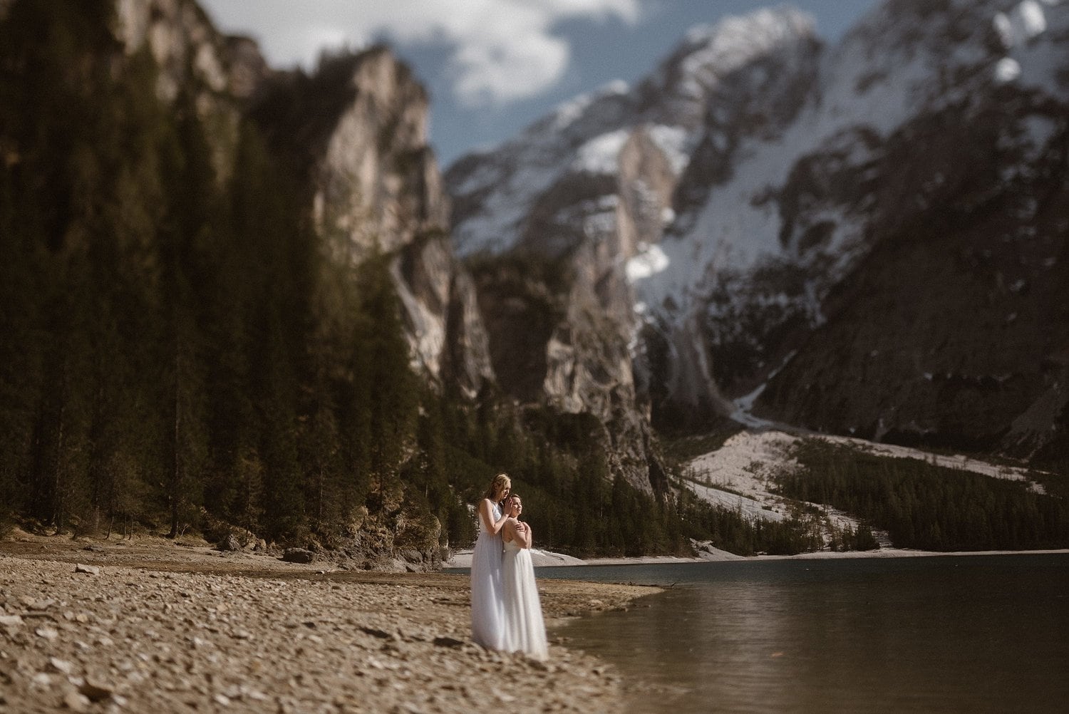 Two brides embrace, while standing on the shore at Lago De Braies in Italy. 