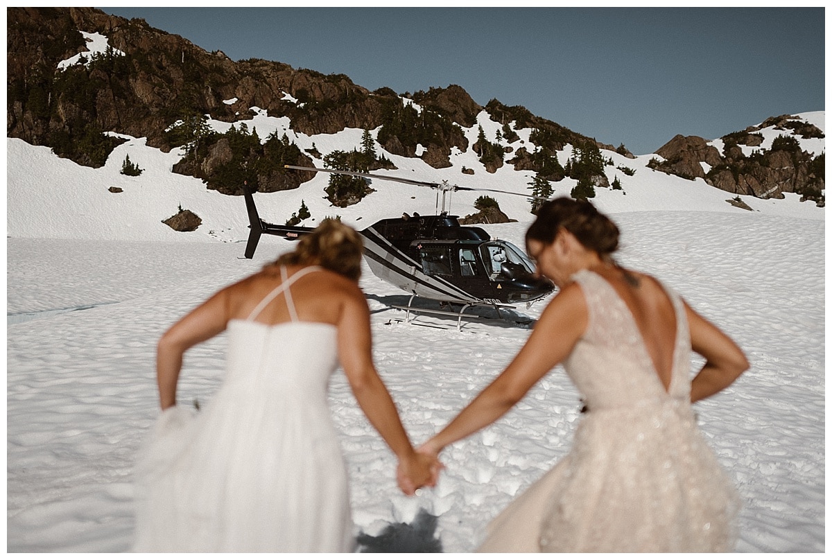 Two brides run through the snow on a glacial mountain in Tofino, Canada. There is snow covering the ground and a helicopter in the background. 