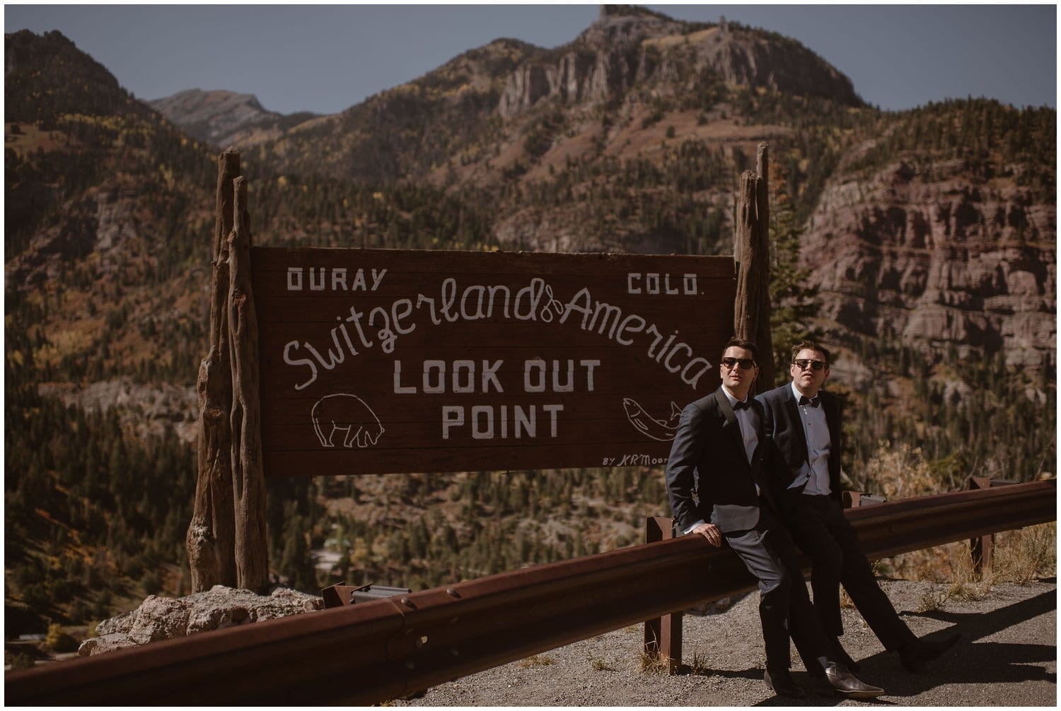 Grooms stand in front of sign at look out point in Ouray, Colorado. 