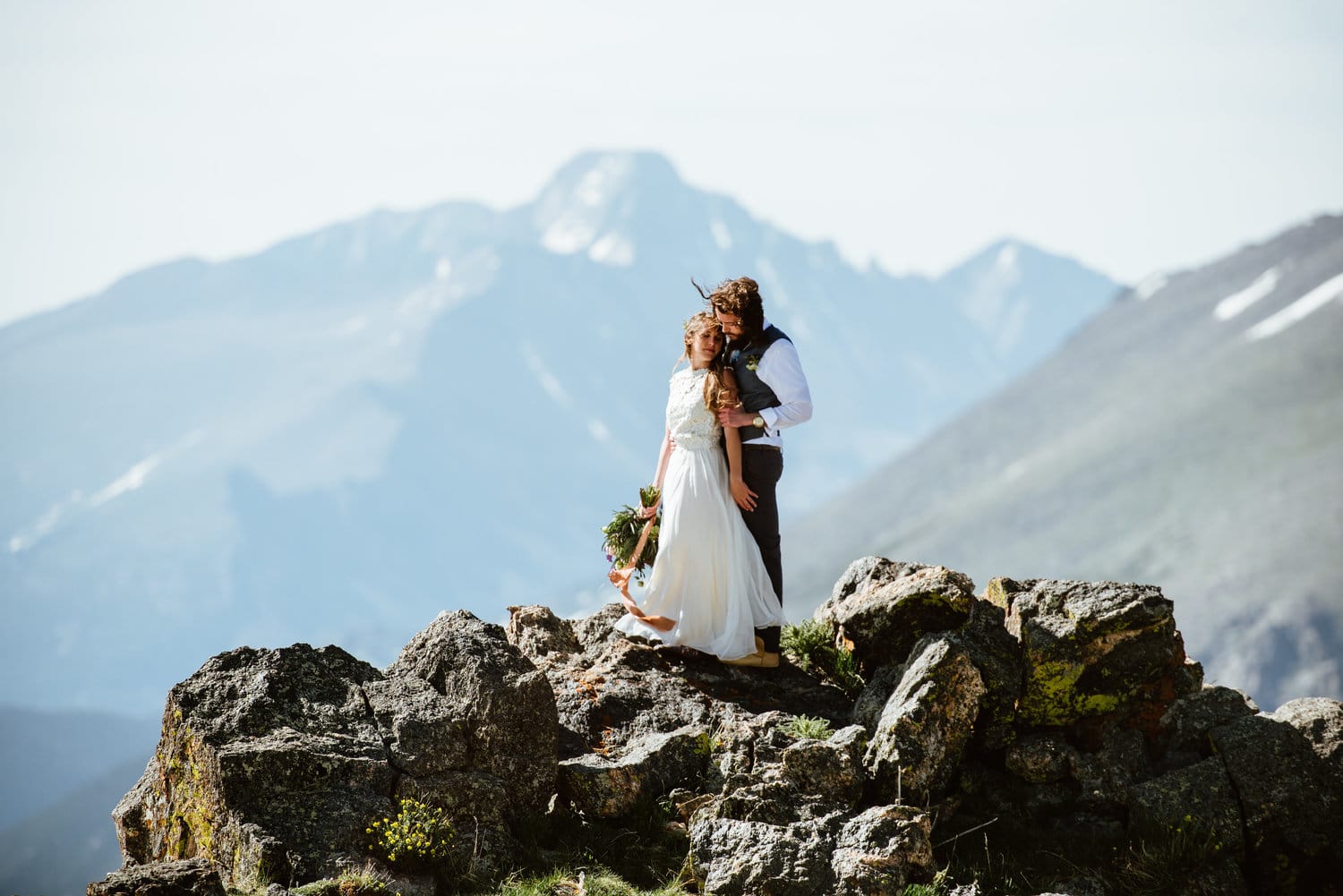 Bride and groom embrace at the top of a cliff on their elopement day at Trail Ridge Road, Colorado. There are mountains in the background. 