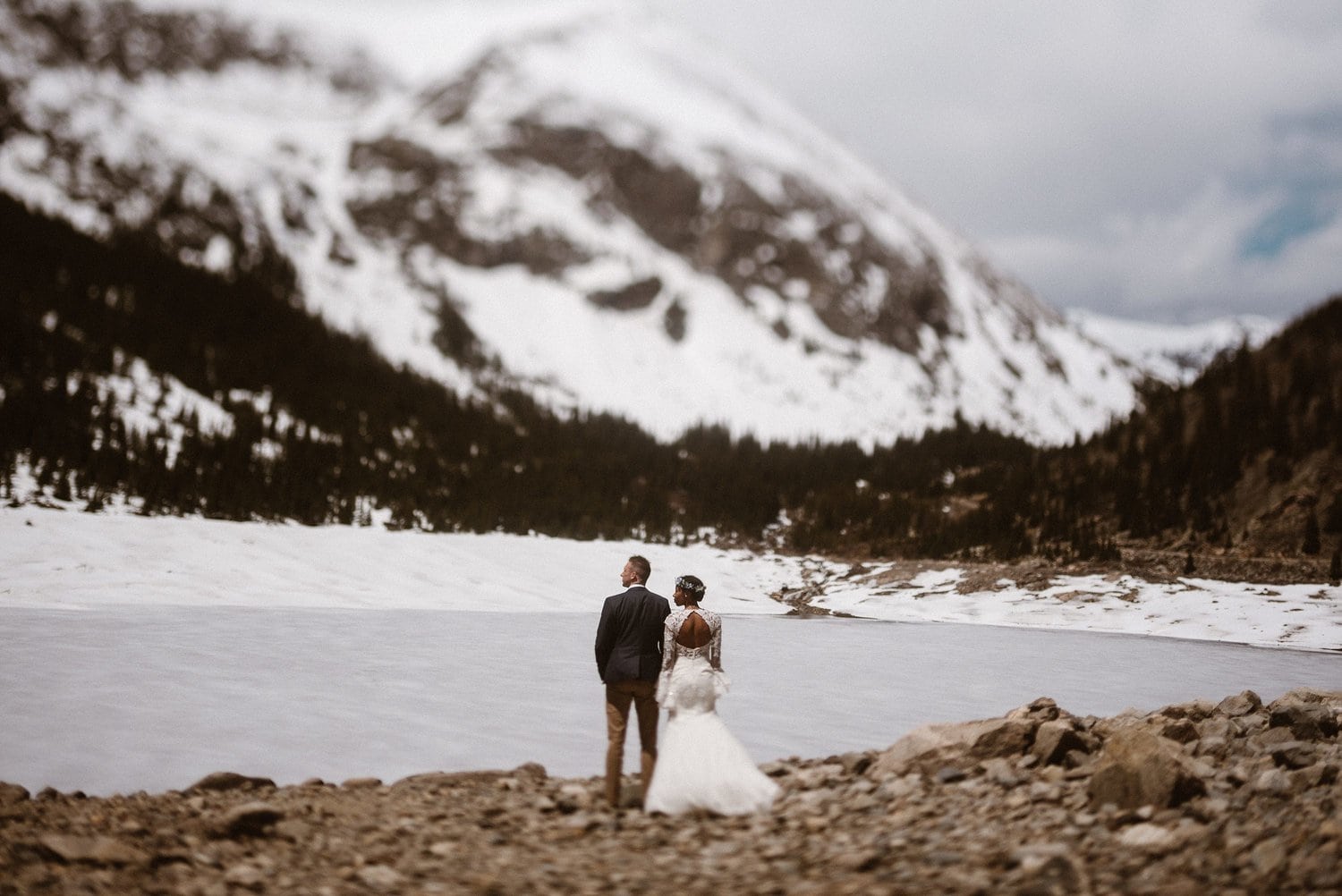 Bride and groom stand together and look out at frozen lake and mountains at Loveland Pass, in Colorado. 
