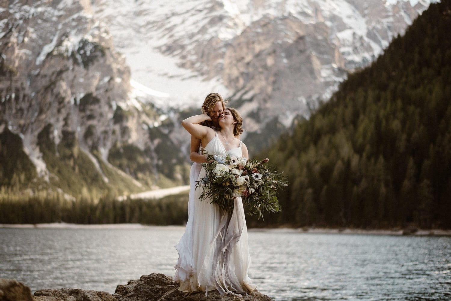 two brides embrace while standing on the shore at Lago De Braies in Italy. 