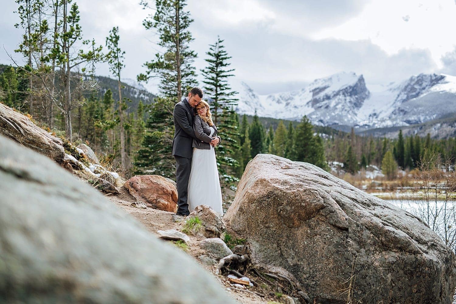 Bride and groom embrace in front of Sprague Lake in Rocky Mountain National Park, Colorado. 