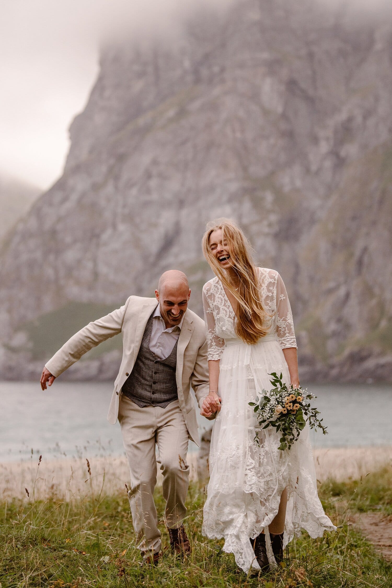 The bride and groom hold hands while laughing together on their elopement day, at a beach in Norway