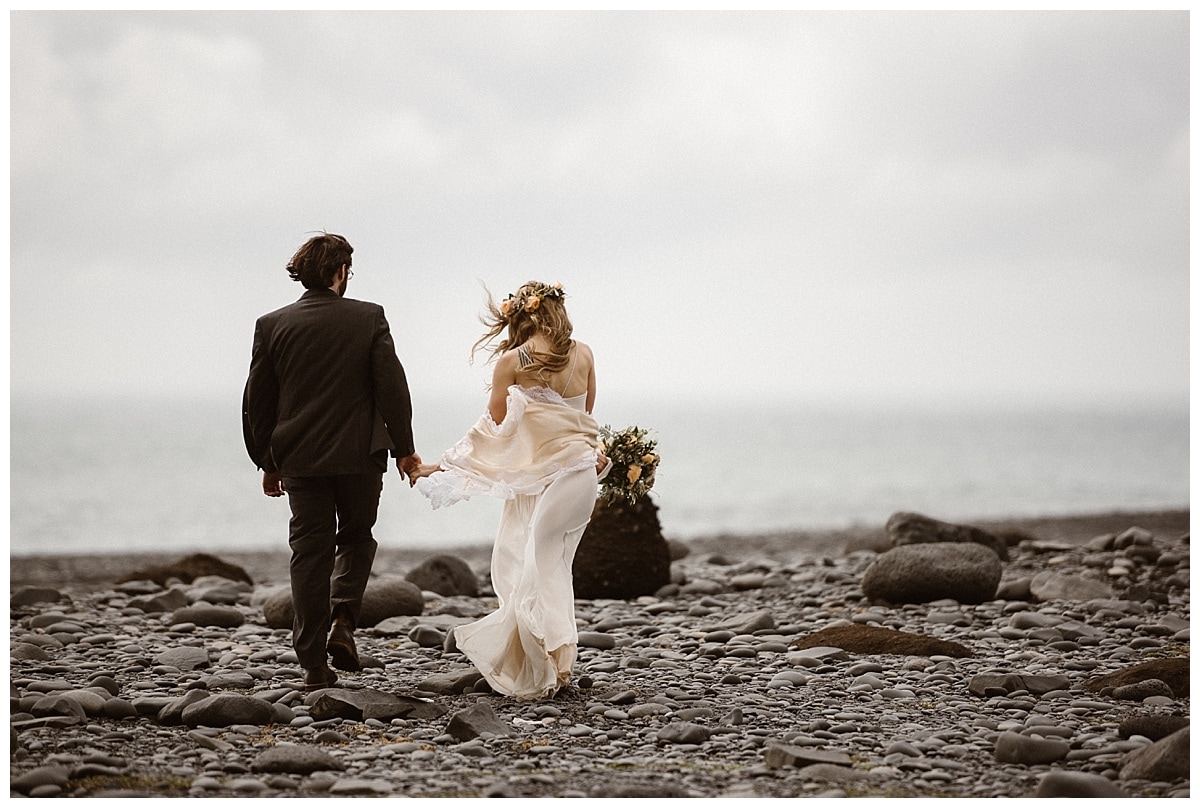 Bride and groom walk along a beach, while holding hands, in Iceland. 