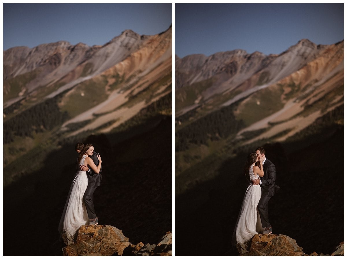 Bride and groom embrace while  standing on a rock together. The brides dress flows in the wind at Ophir Pass, in Colorado. There are mountains in the background. 