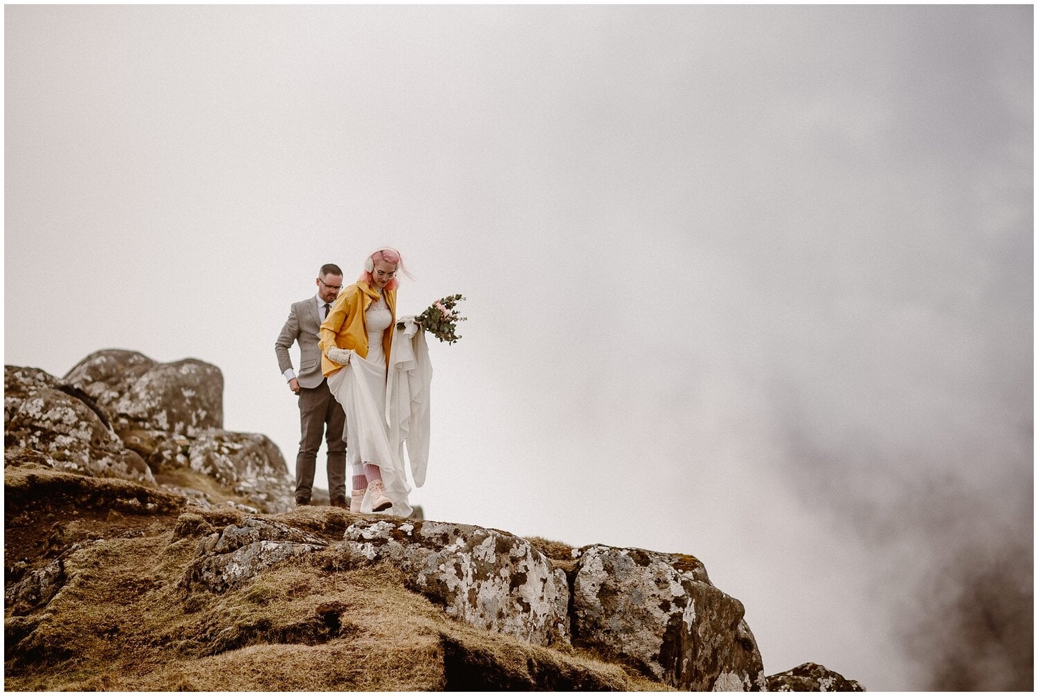Bride and groom hike up mountain with fog in the background. 