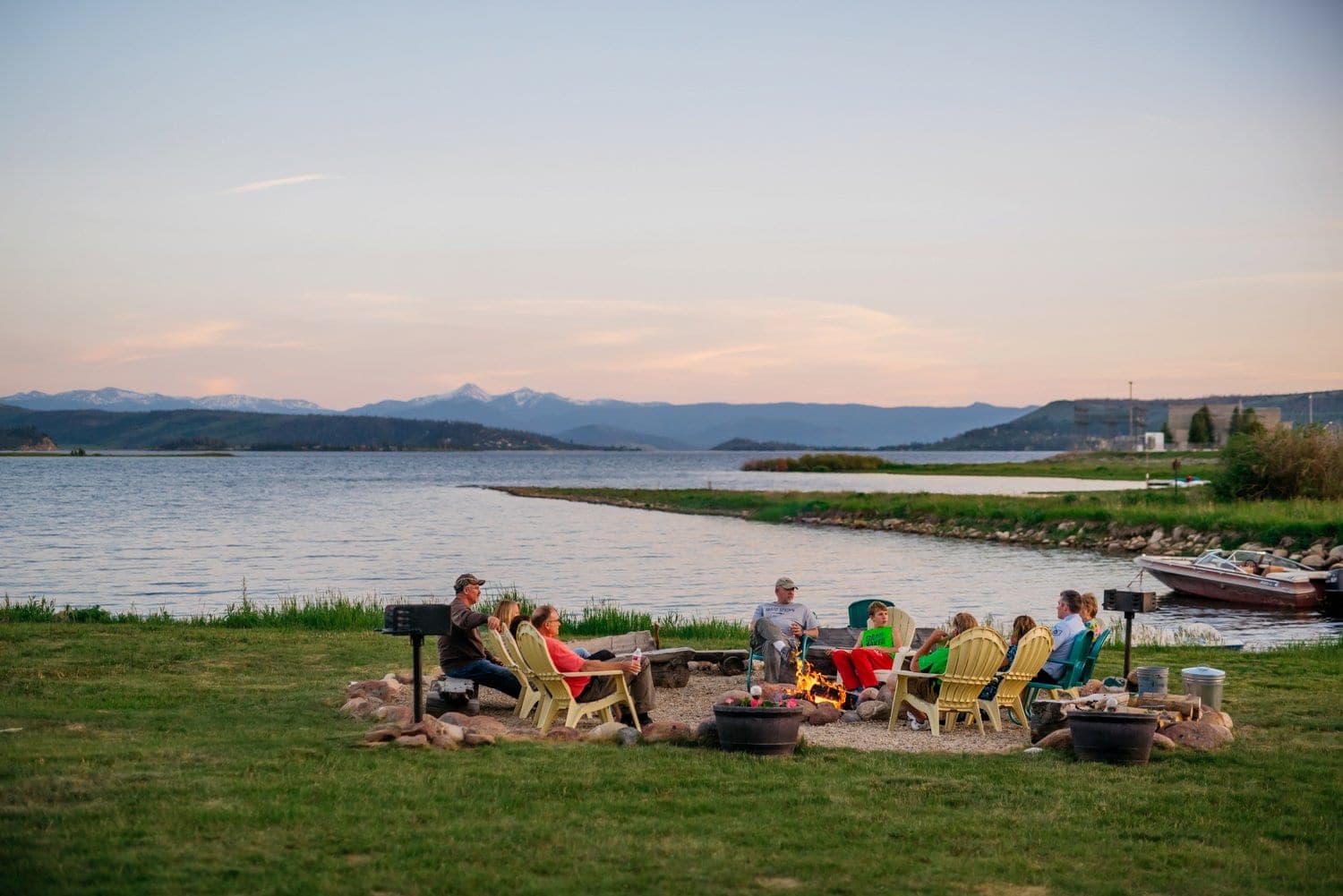 Friends and family sit in front of a campfire in Grand Lake, Colorado, There is a lake and mountains in the background. 