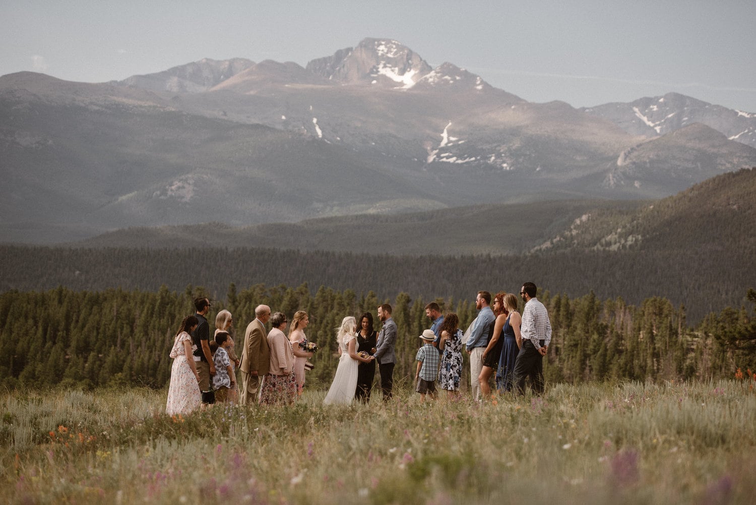 Bride and groom are surrounded by friends and family during their intimate elopement ceremony. They are standing in a meadow with wildflowers. There are trees and mountains in the background. 