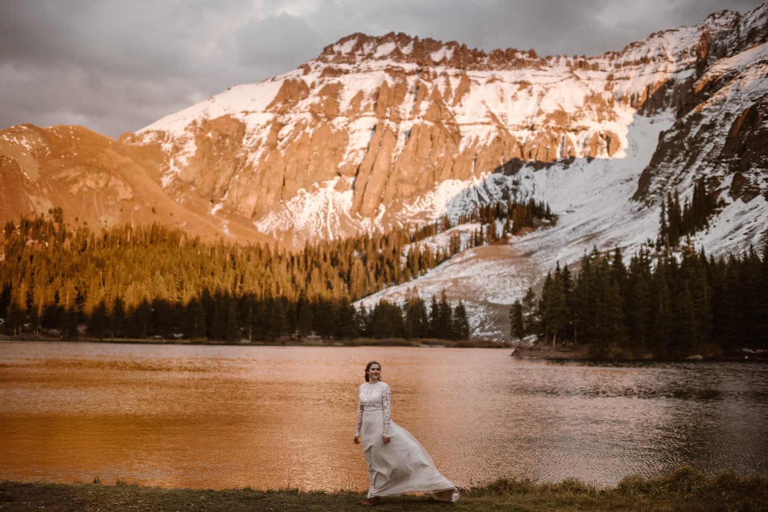Bride wearing a white dress and standing in front of an alpine lake. There are snow-capped mountains with alpenglow in the background. 
