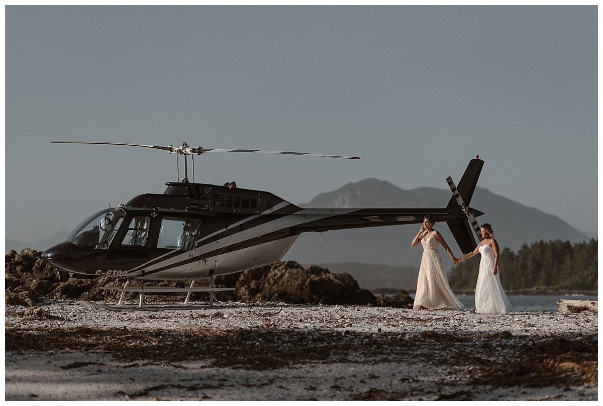 Two brides hold hands while standing in front of a helicopter on Shell Island in Tofino, Canada. 